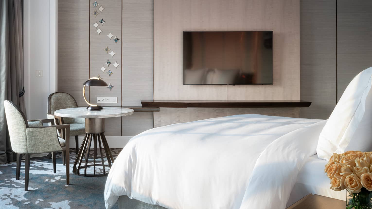 A modern bedroom featuring a large bed with white linens, wall-mounted TV, small round table with chairs, and decorative wall with metallic accents.