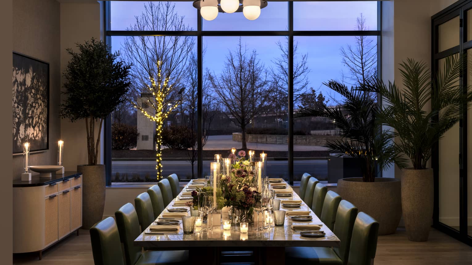 Dining table with candles and place settings, three large plants, and floor to ceiling windows.