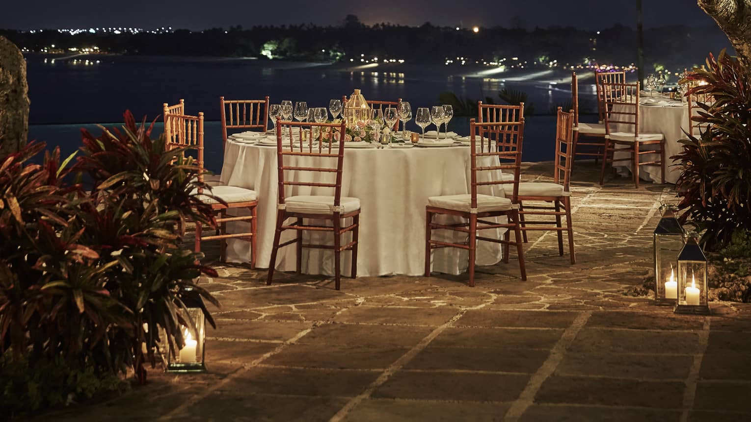 A romantic dinner table set outside under the night sky, with string lights and lanterns hanging in the trees above it