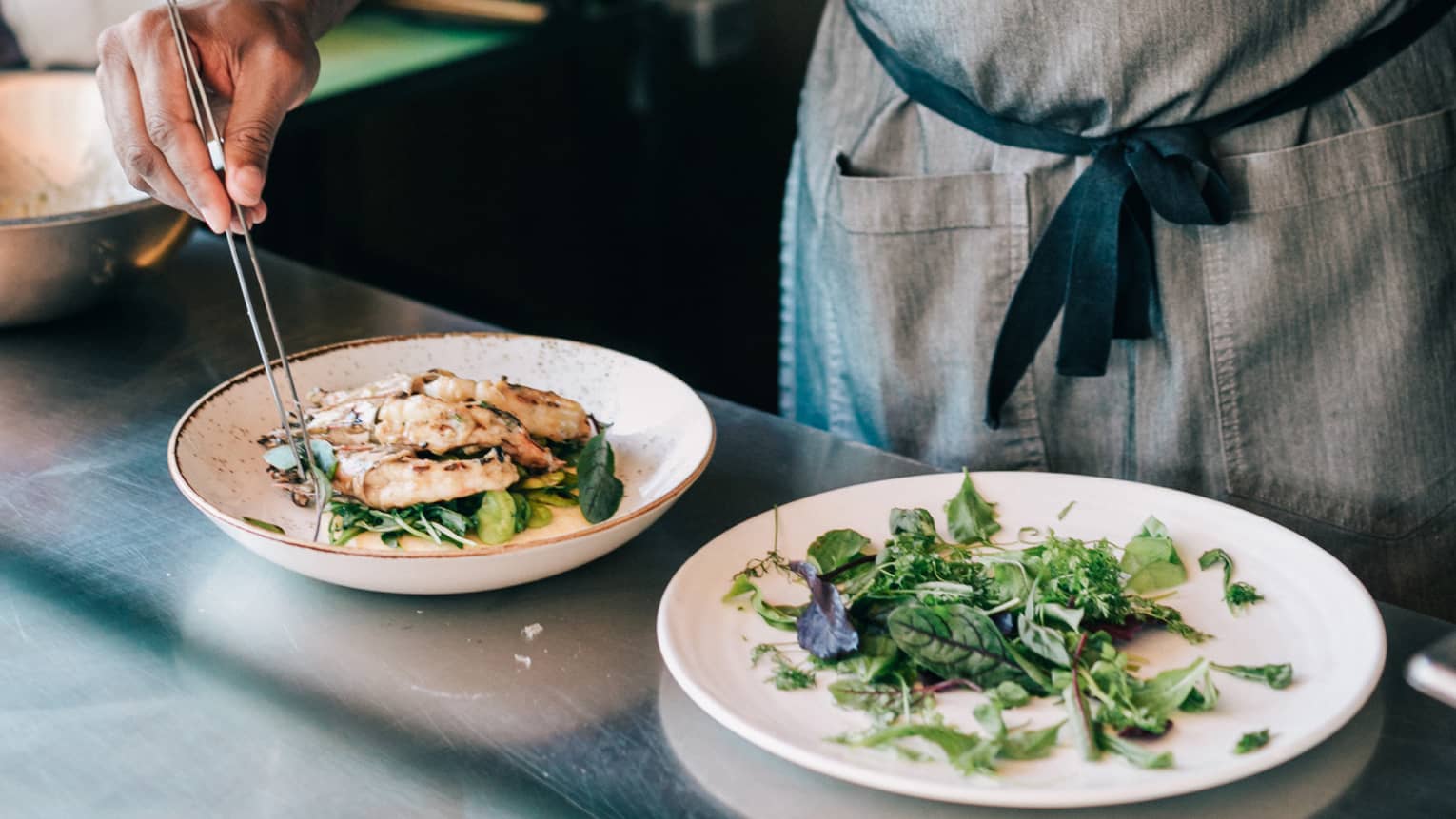 Chef wearing apron garnishing seafood dish with greens, beside salad plate