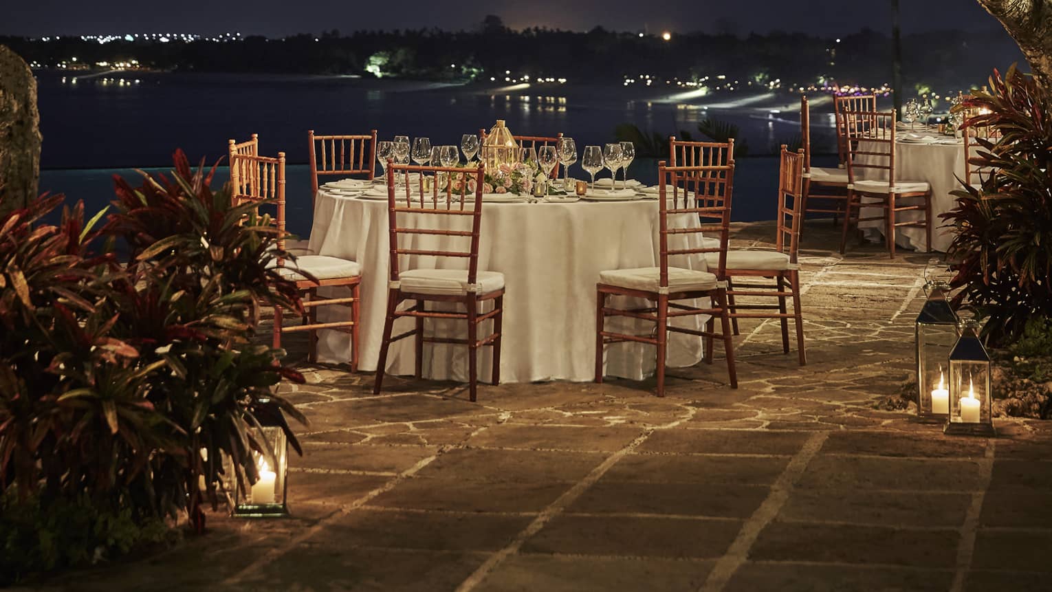 A romantic dinner table set outside under the night sky, with string lights and lanterns hanging in the trees above it