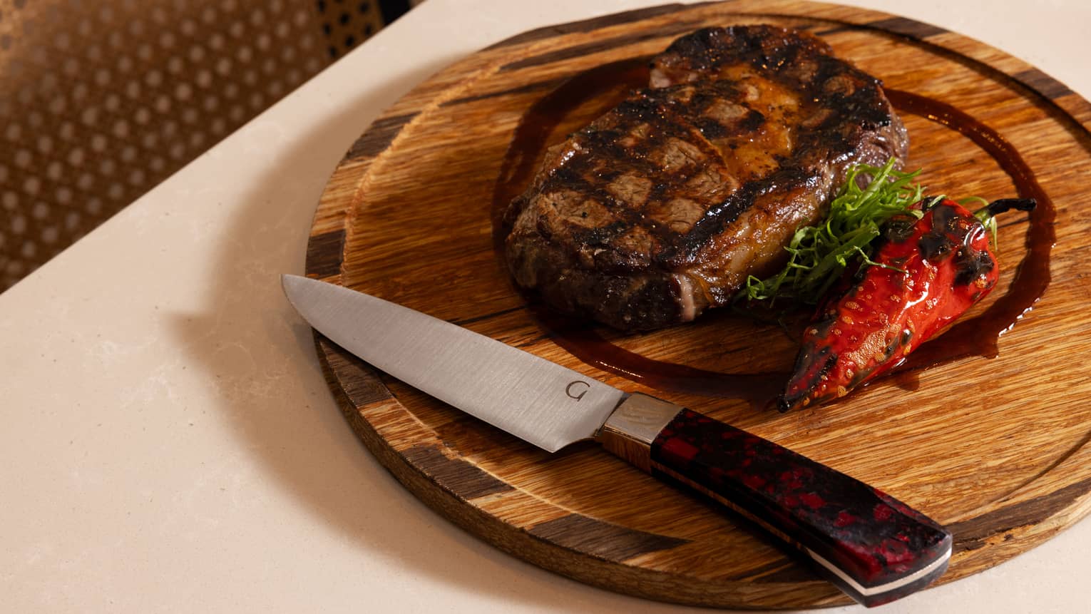 A round wooden serving plate with a piece of steak and garnishes. A steak knife sits on the plate near the edge.