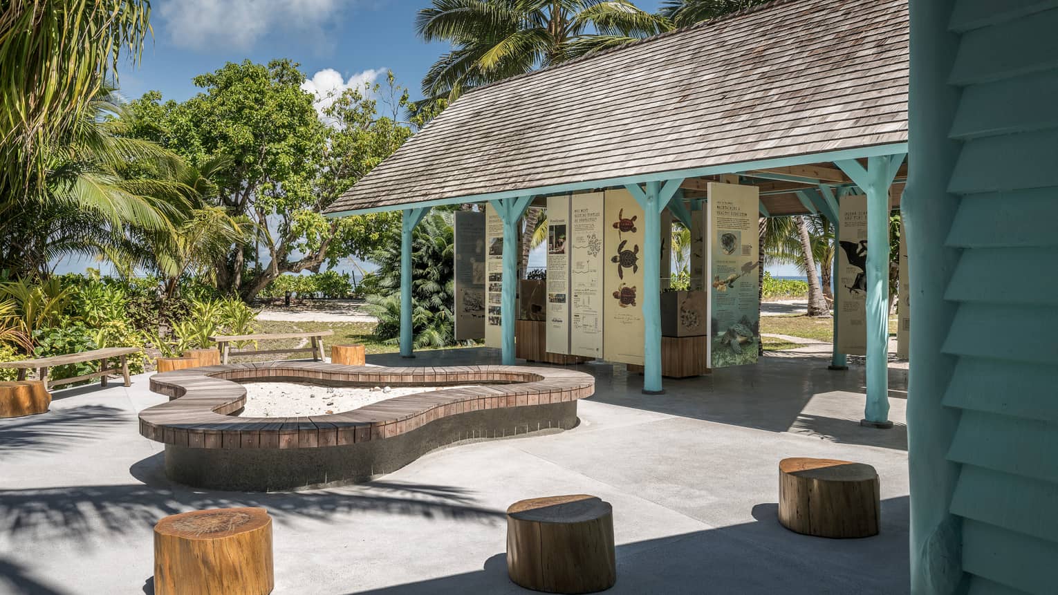 Outdoor information hut with blue poles, surrounded by palm trees, seating made of tree stumps