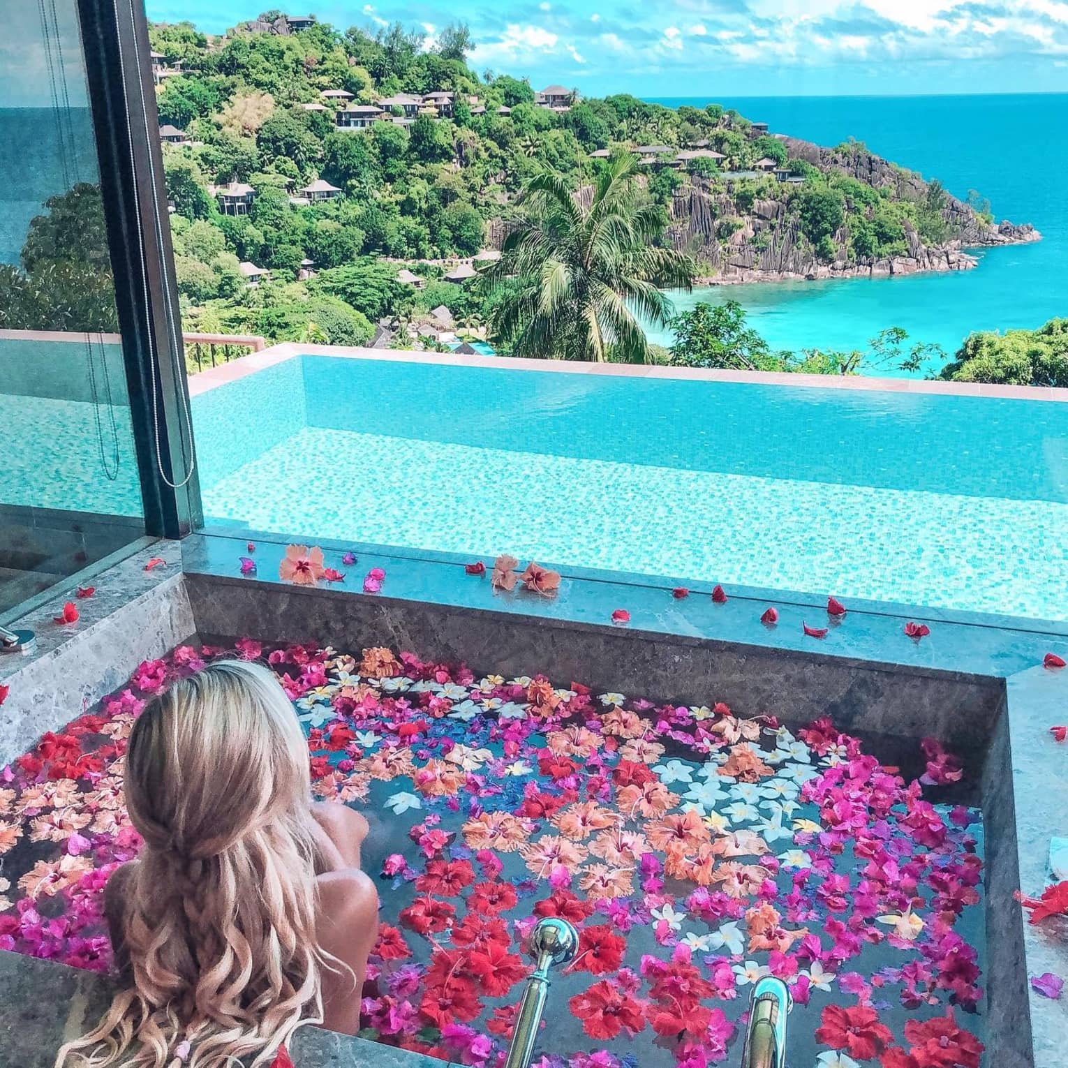 A woman sits in a bathtub full of flower petals by an open window overlooking a private pool, blue waters and a view of the island