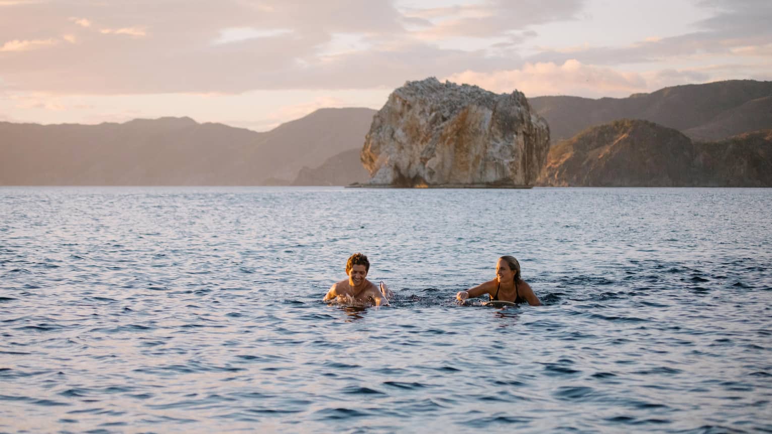 Two people paddling through the water on their surfboards with rock formations in the background and a hazy pink sky