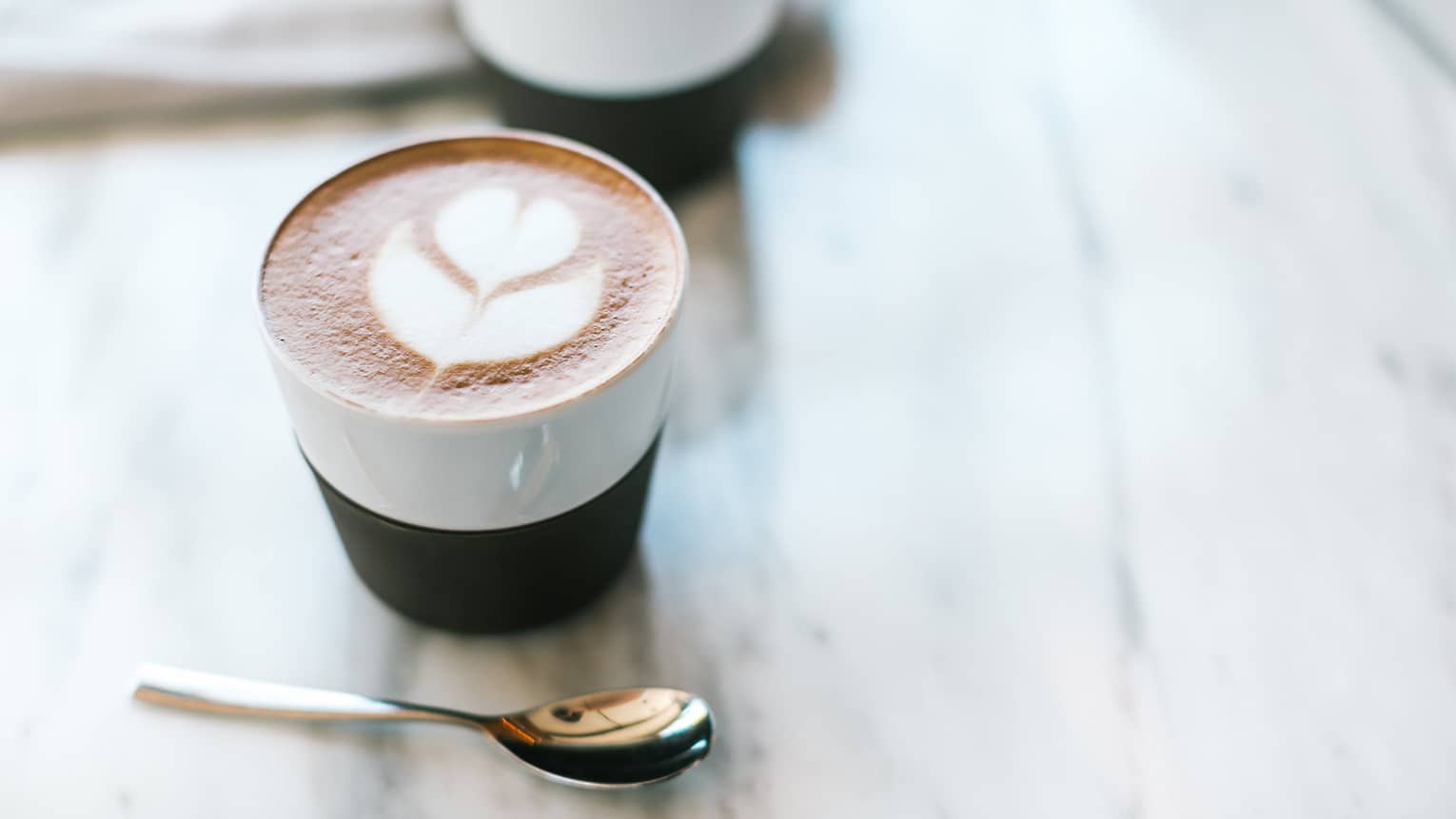 A small cup of coffee with a flower design in the foam next to a small spoon.