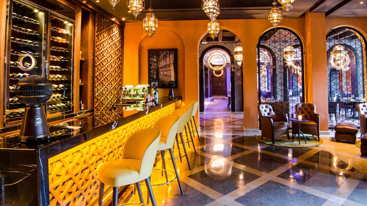 Wine bottles behind glass display in front of decorative carved bar with yellow lights, stools