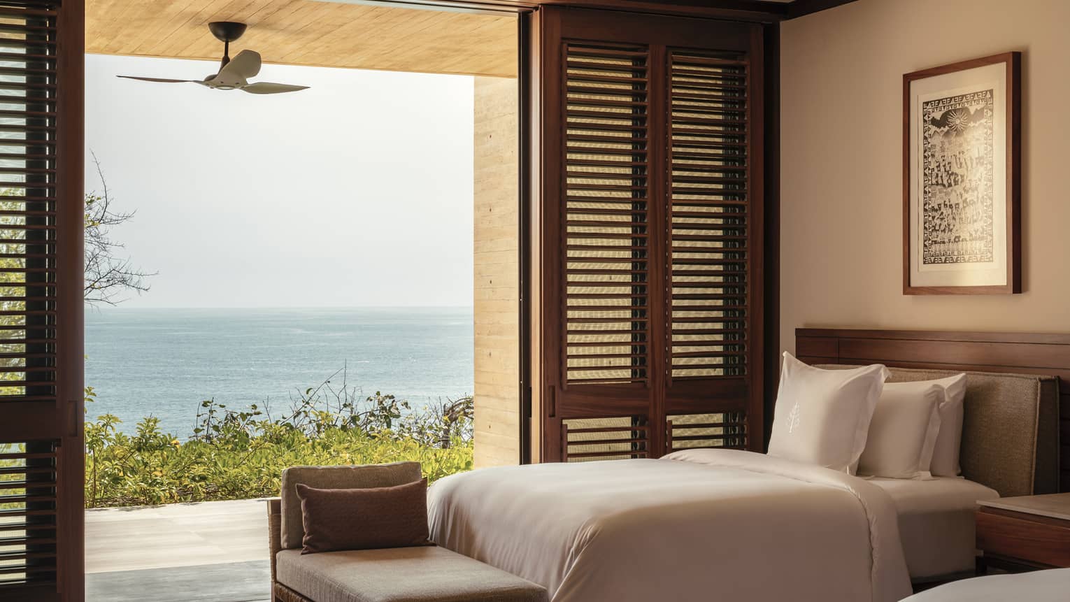 Bedroom with twin beds and white linens, opening to a terrace with ocean views. Wooden shutters and a ceiling fan enhance the room’s airy, tropical feel.