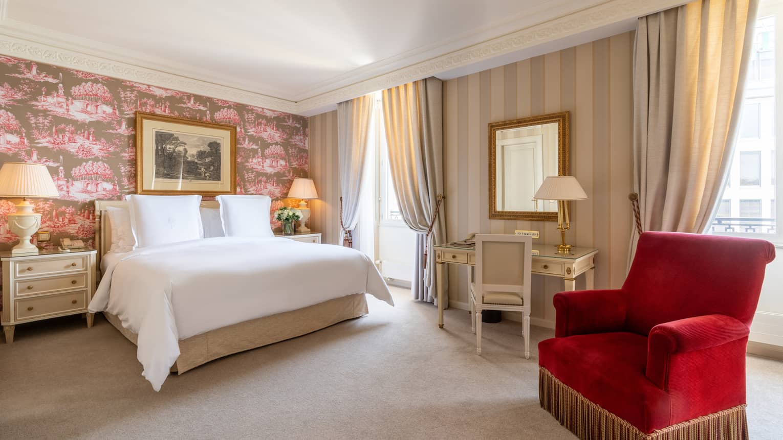 Bedroom with white bed, flanked by nightstands against toile-papered wall with painting, desk and red club chair