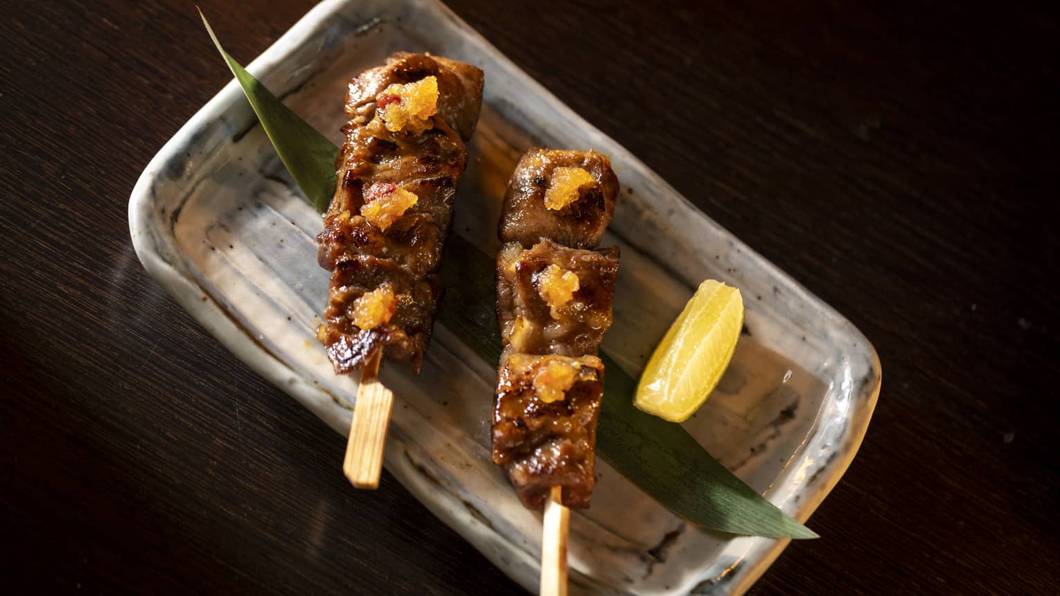 An artistic rectangular glazed plate holds skewers of food with garnishes.