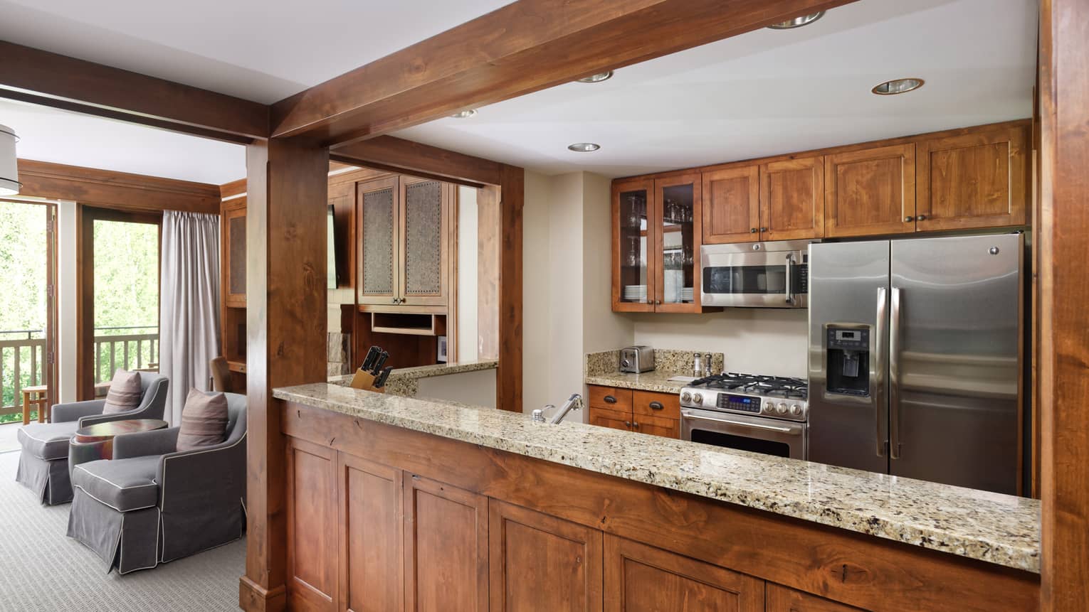 Kitchen with wooden cabinets, stainless steel appliances and granite countertops, opening to a living room with two grey armchairs