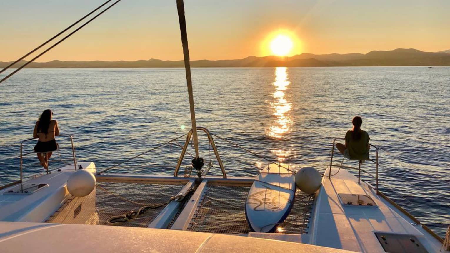 Two guests lounge on luxury sailboat near stern railing at sunset