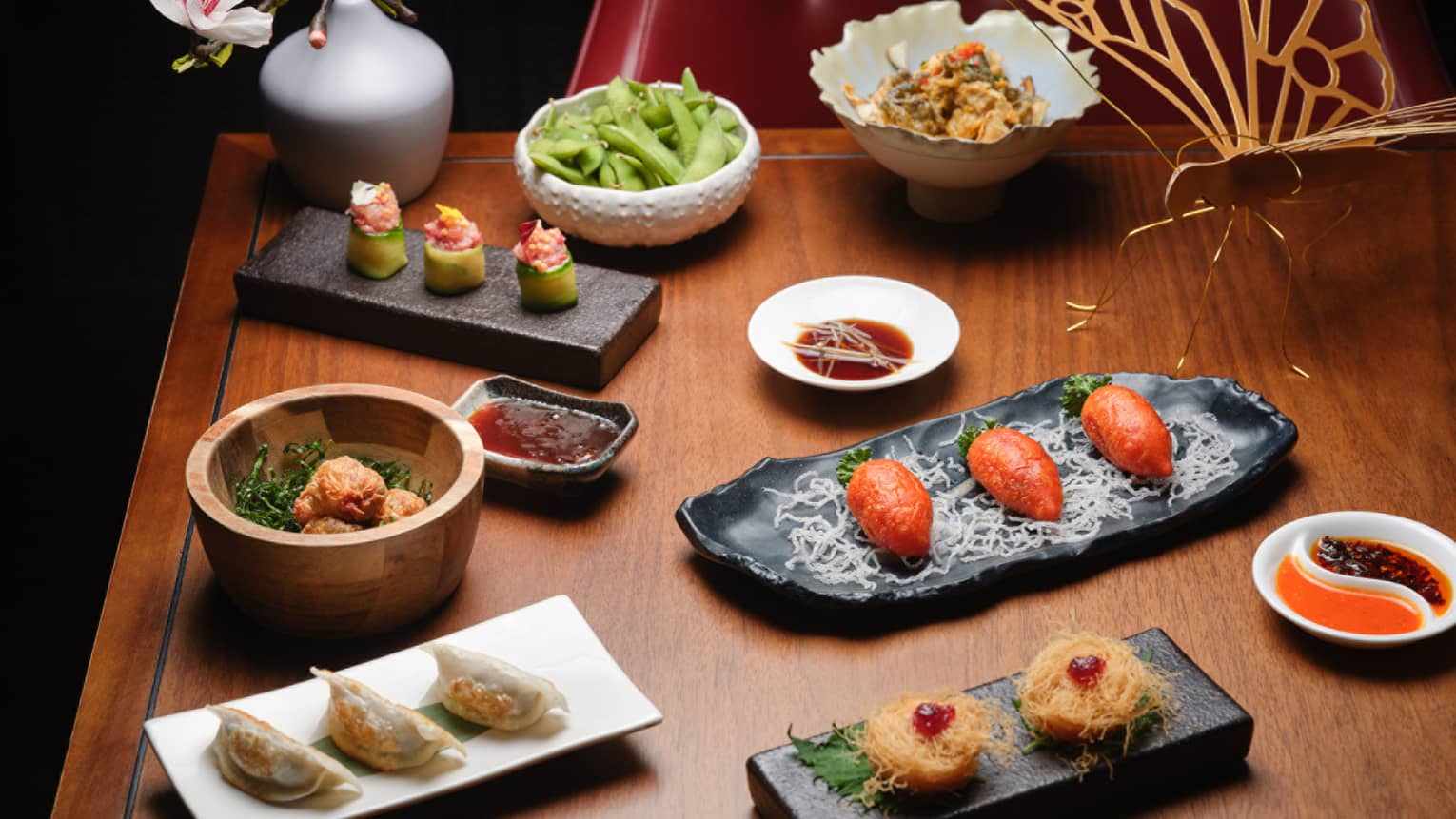 An array of plates and bowls in various sizes and shapes contain colourful food items and are set on a wood table. A delicate cut-out butterfly sculpture and a floral arrangement sit next to the dishes.