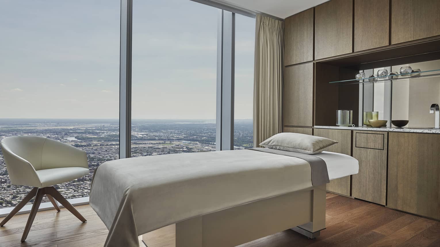 A massage platform with a sheet and small pillow next to large windows, a small white chair, cabinets and a shelf.