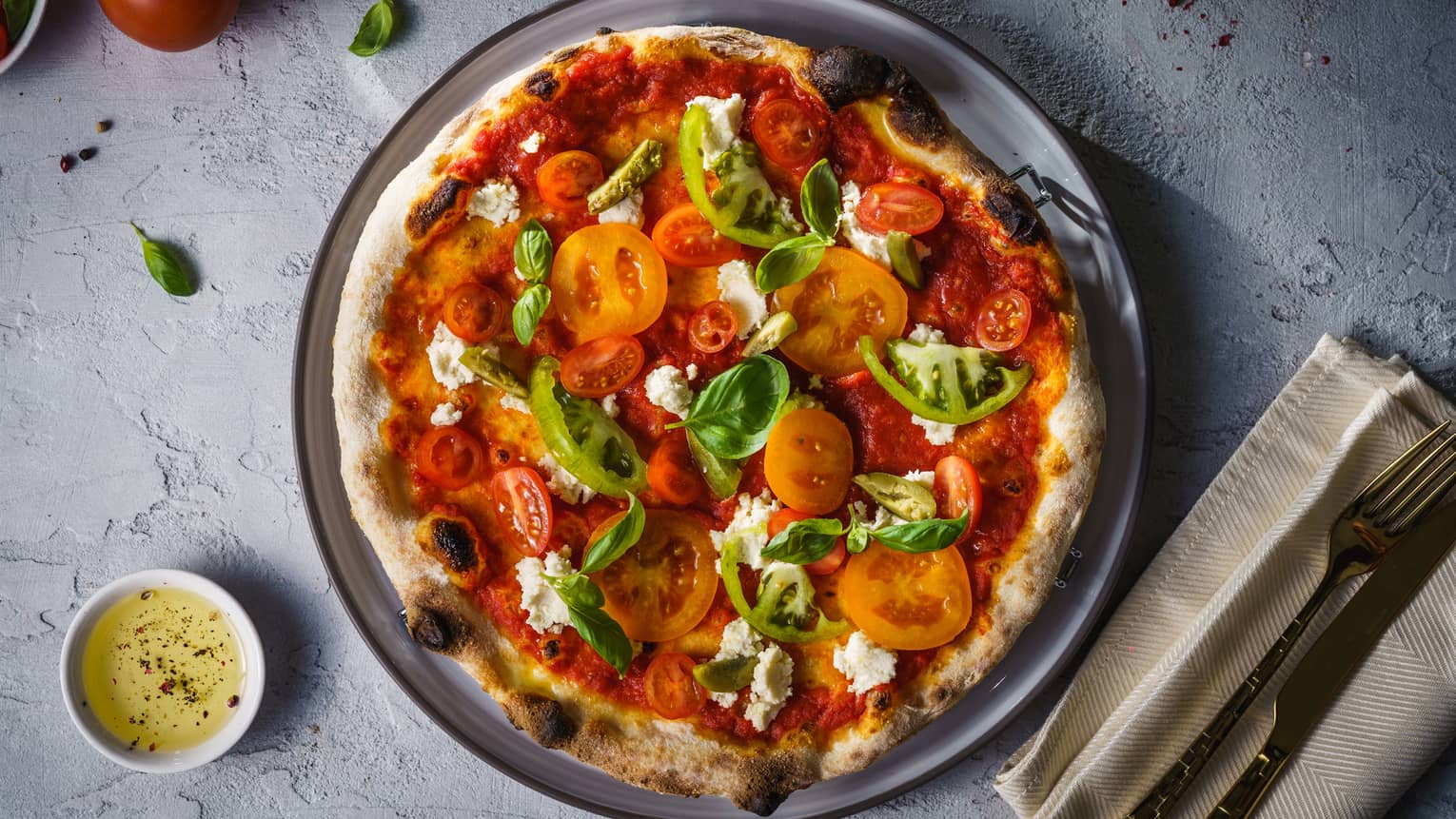 A pizza covered in tomatoes, peppers and spinach served on a grey plate.