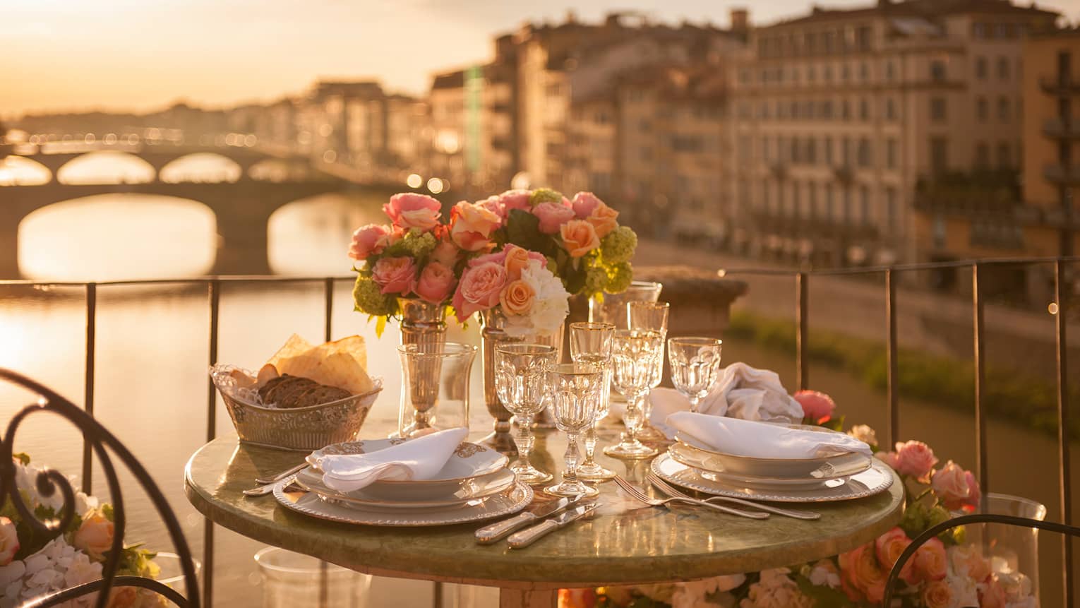 A romantic table for two is set with white porcelain plates, crystal glasses, pink roses on a balcony overlooking a bridge and historic building in Florence