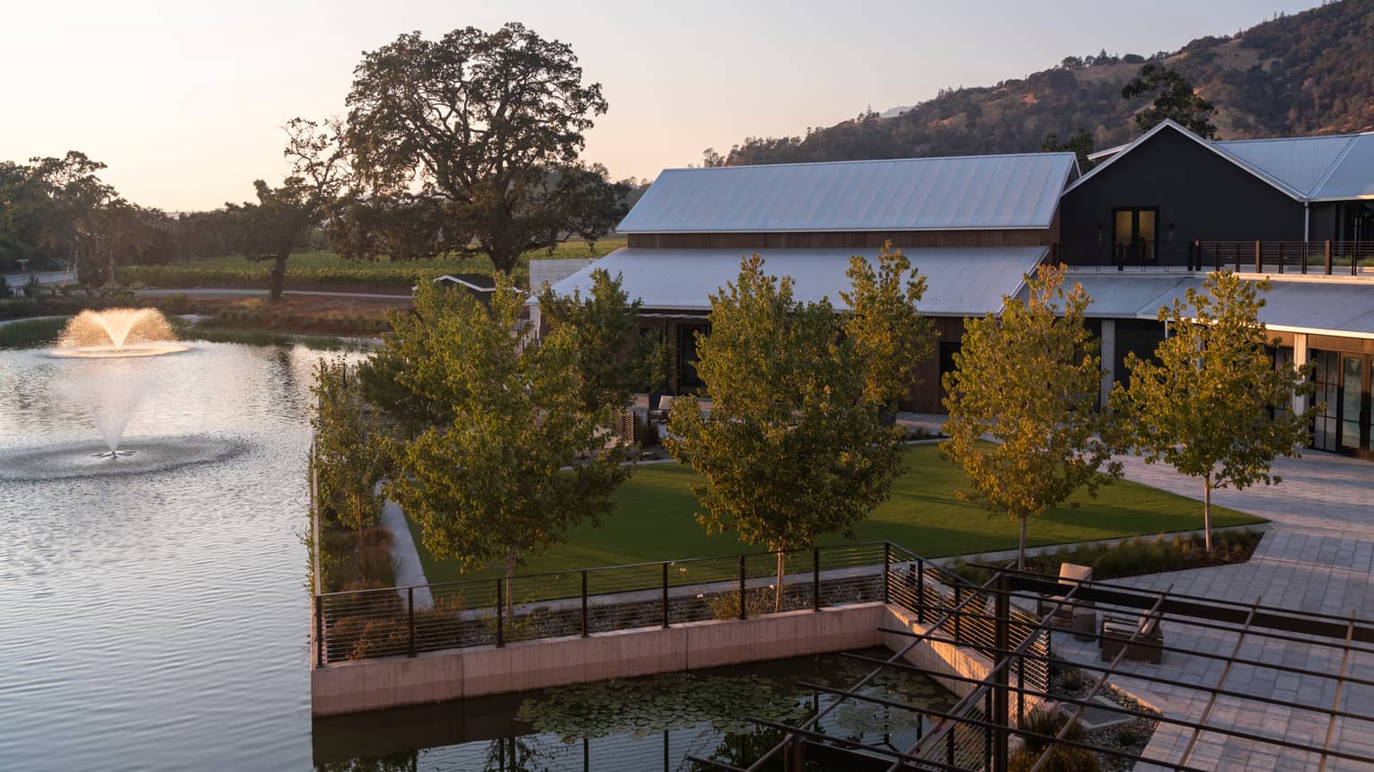 A spacious green lawn space surrounded by trees, a seating area and a lake with a fountain