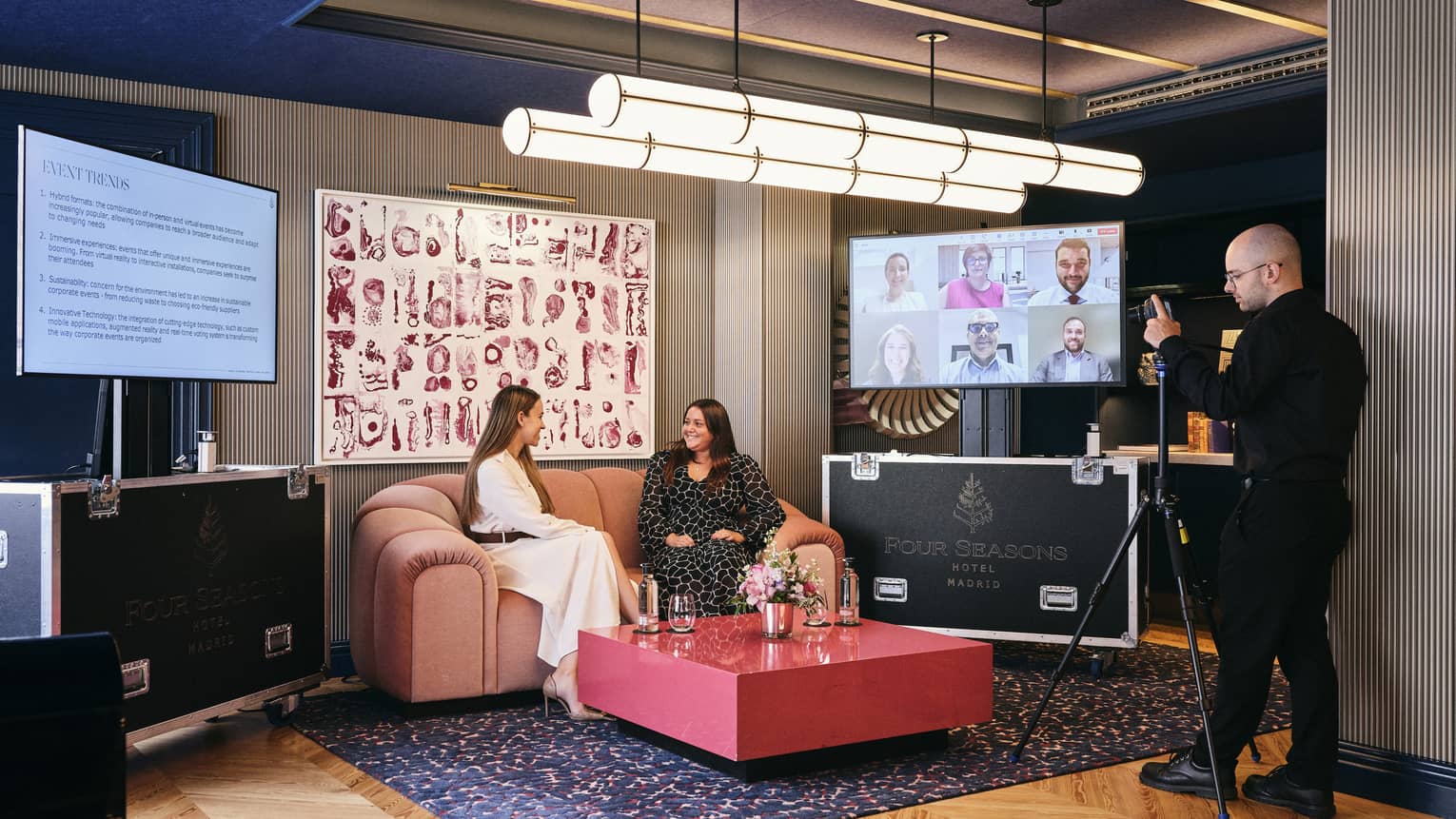 Two people sit on a couch in a well-lit meeting space with wood flooring and an area rug. Another person stands in front of them with a camera on a tripod. Art hangs on the wall behind the couch, and the couch is flanked by two flat-screen monitors. A coffee table is in front of the couch.