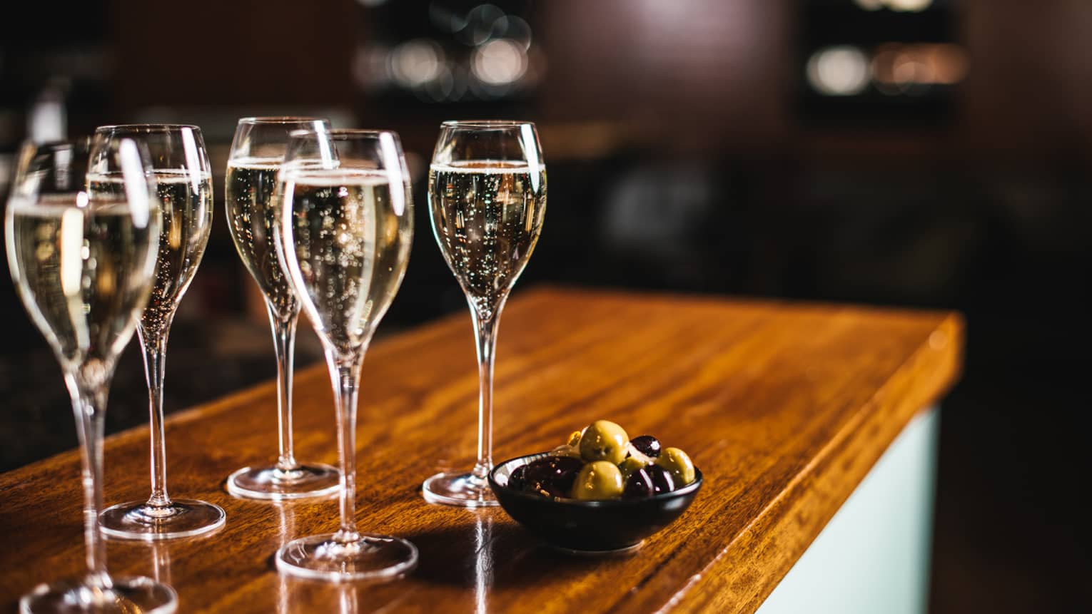 Five glasses of sparkling wine with bowl of black and green olives, atop a wooden bar top