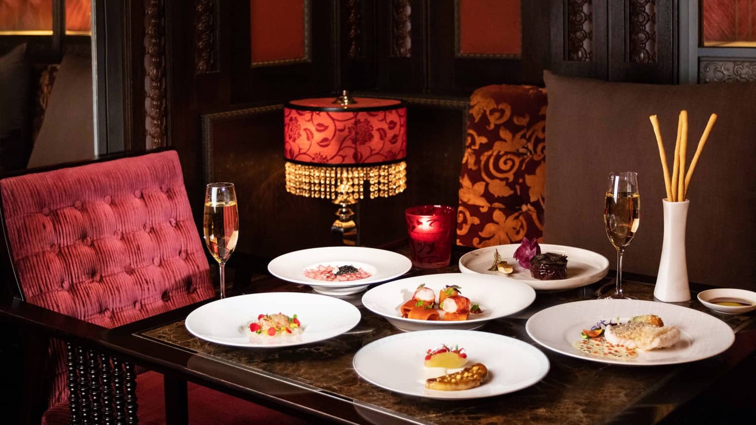 A dining table with maroon upholstered seating, with various colourful culinary dishes served on white plates and two glasses of champagne