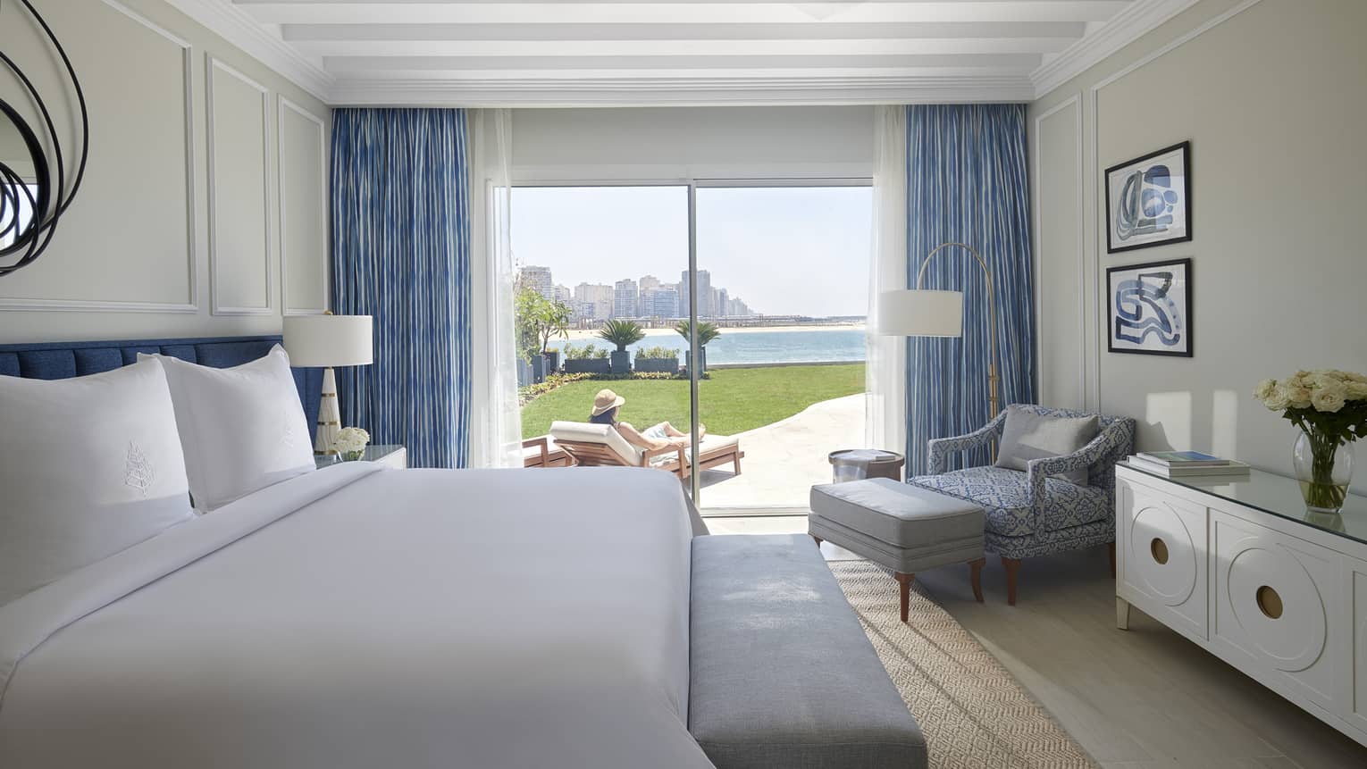 Hotel guest room bed, dresser and chaise to patio door where woman reads in sun