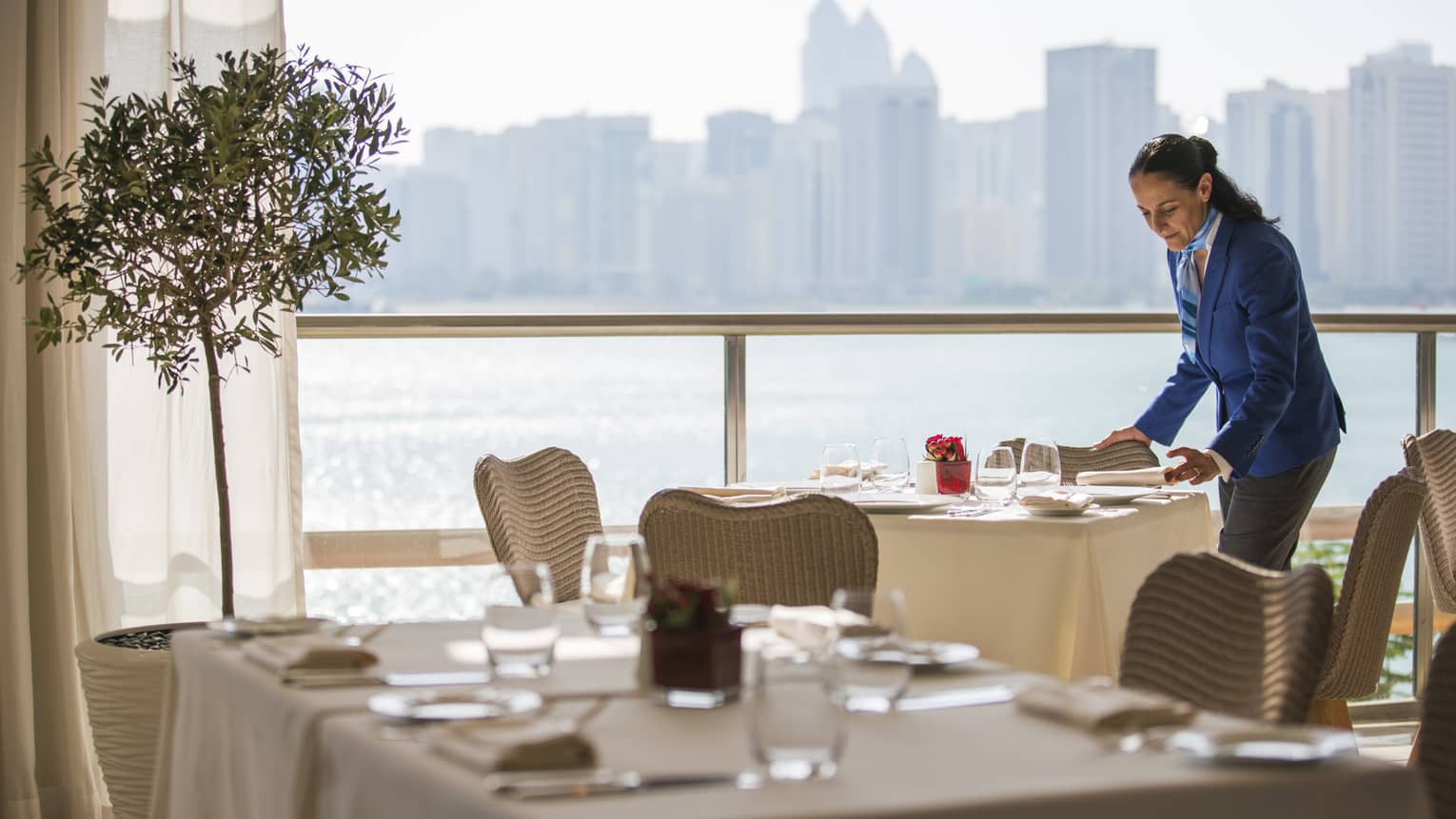Woman in blue suit sets table by sunny window with water views at Cafe Milano