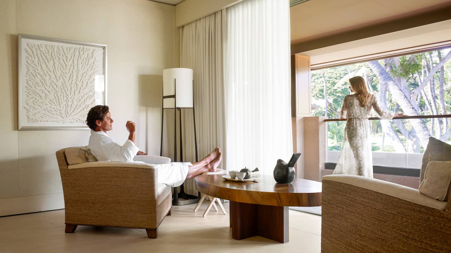 Man in white bathrobe kicks back in armchair in Spa room, woman in sheer robe stands at sunny window