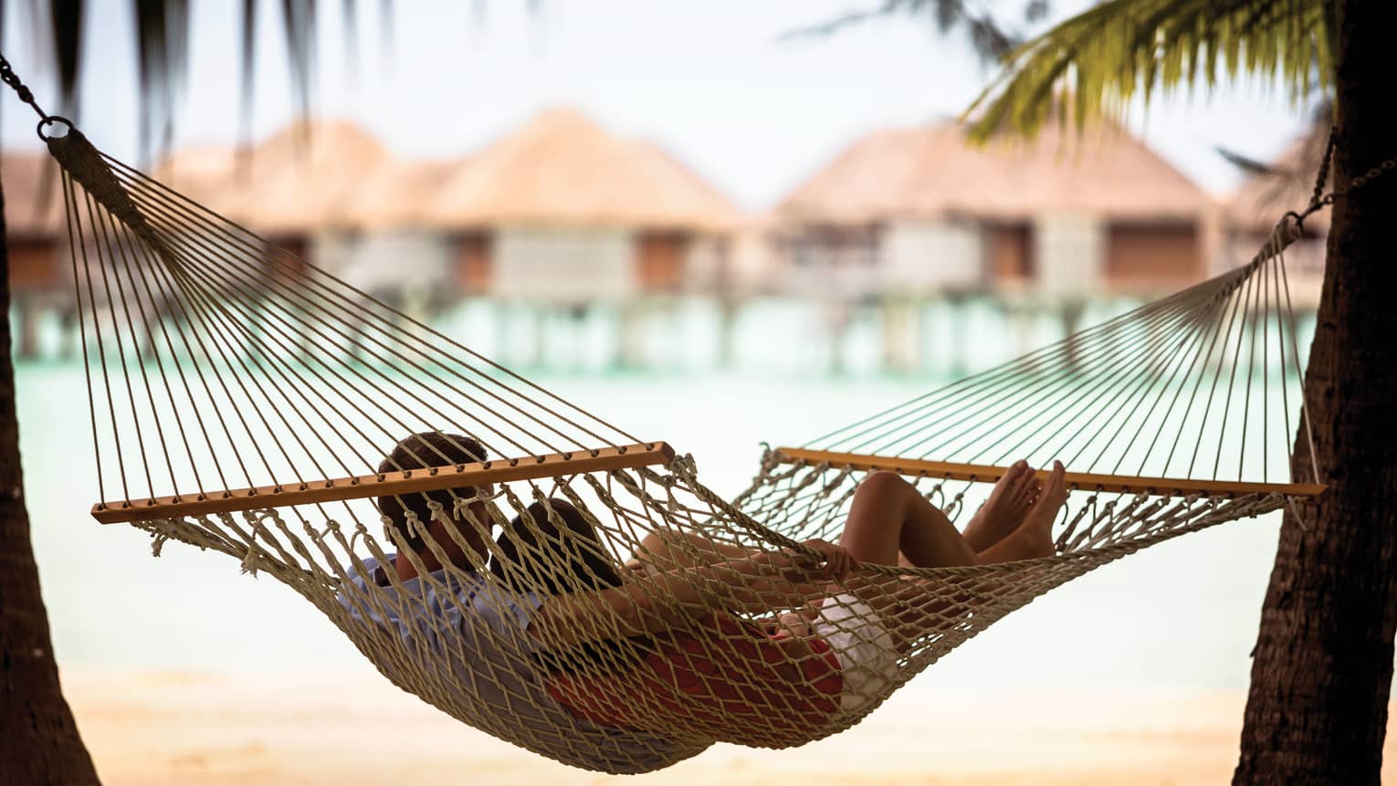 Couple lies in rope hammock between two palm trees, with beach, ocean and overwater bungalows in background