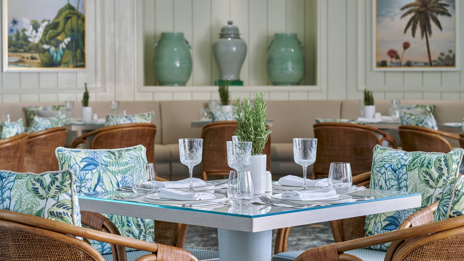 Byblos indoor dining area with rattan chairs and blue patterned accent pillows, square glass-topped tables