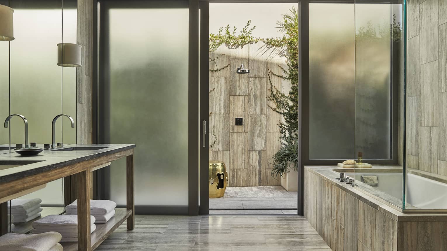 Modern bathroom with dual sinks, a soaking tub, frosted glass doors and an outdoor stone shower with greenery and a golden accent stool