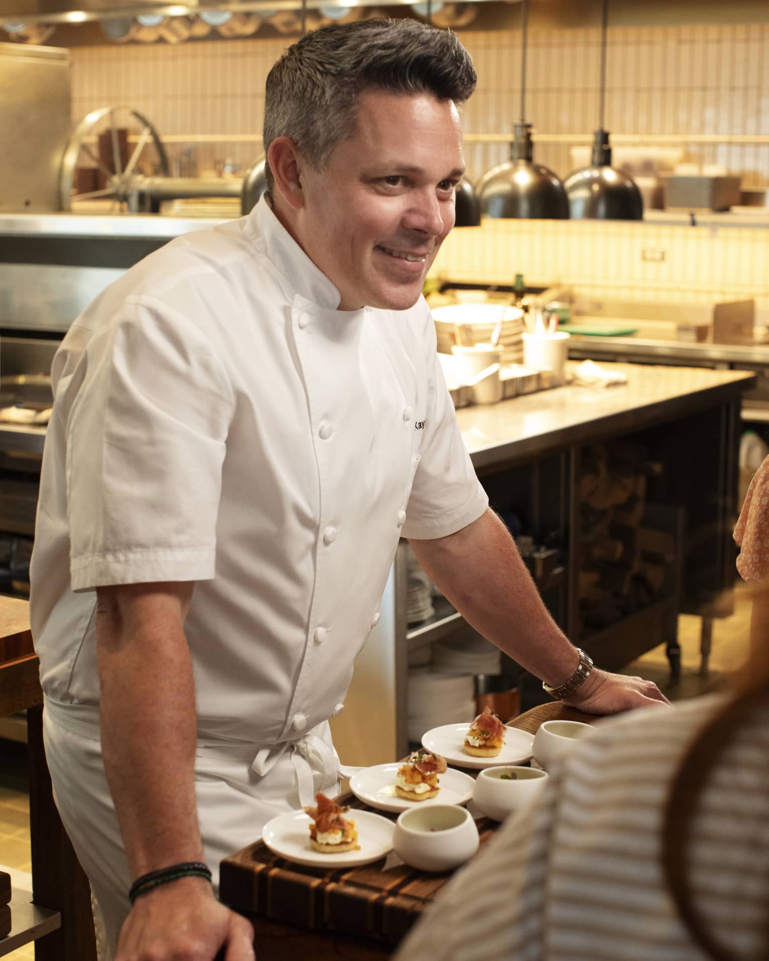 Chef standing beside a dining table in the kitchen talking to guests