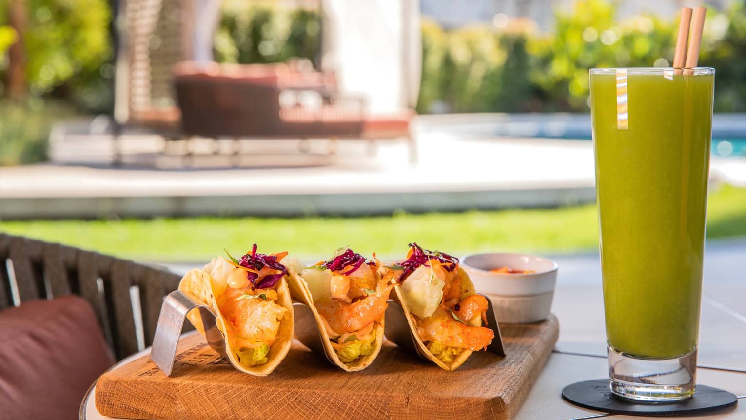 Poolside table set with three shrimp tacos on board beside tall green cocktail