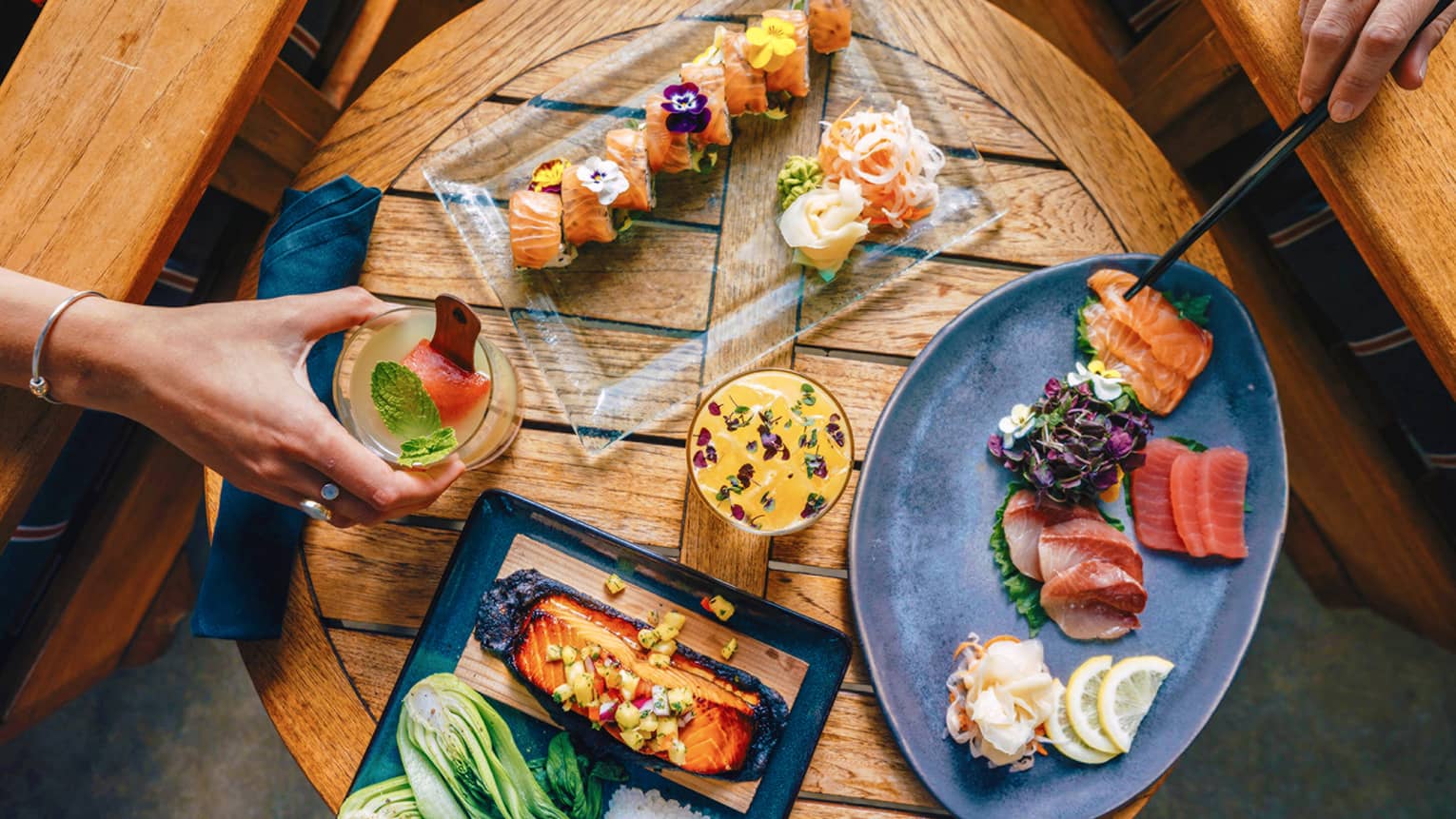 An aerial view of guests enjoying sushi and cocktails on a small, round wooden table 
