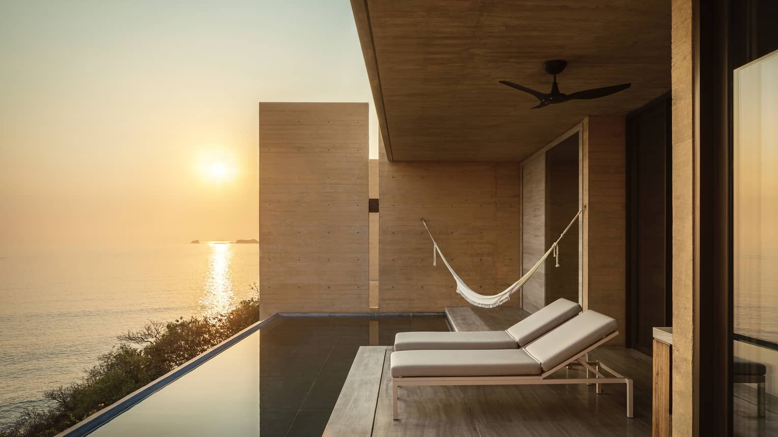 Modern terrace with lounge chairs, a hammock and an infinity pool, overlooking a serene ocean at sunset. The space features minimalist wooden walls and peaceful ambience.