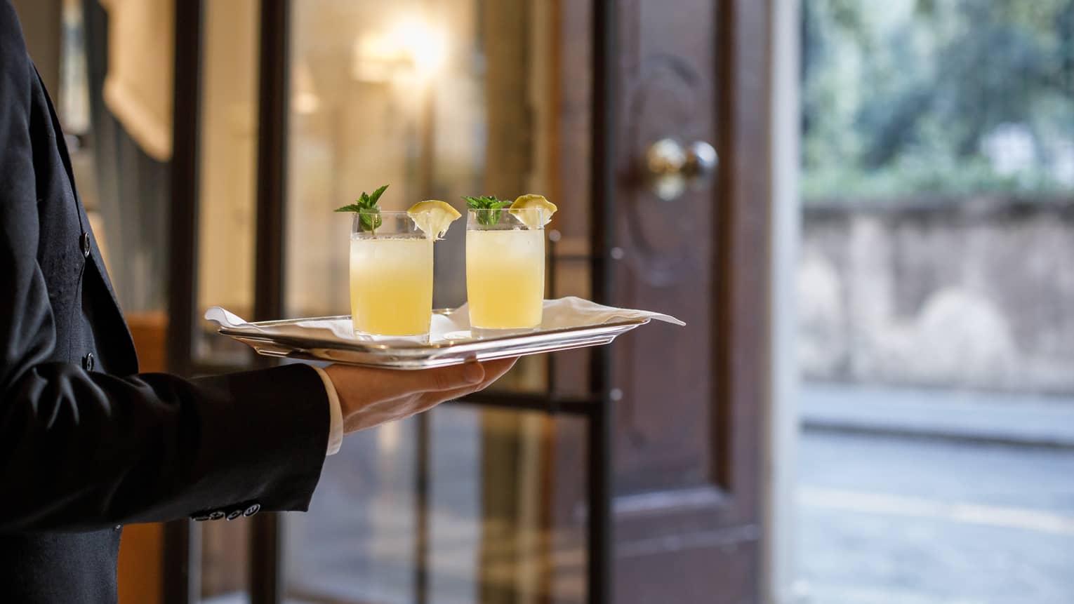 Close-up of server holding tray with two yellow cocktails with lemon, mint garnish