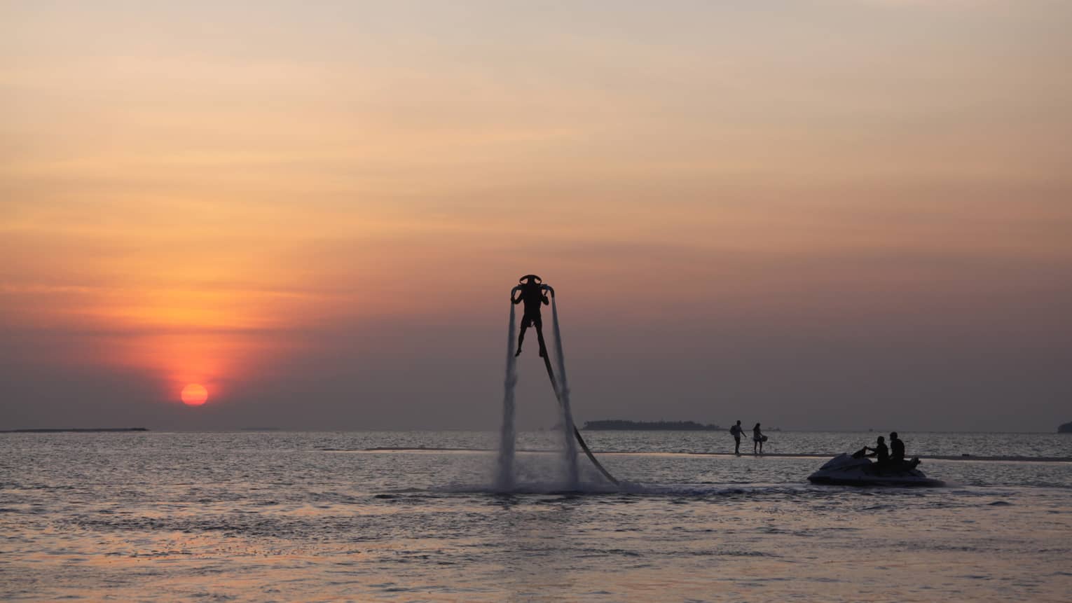 Silhouette of someone with X Jetpack suspended in air over water at sunset
