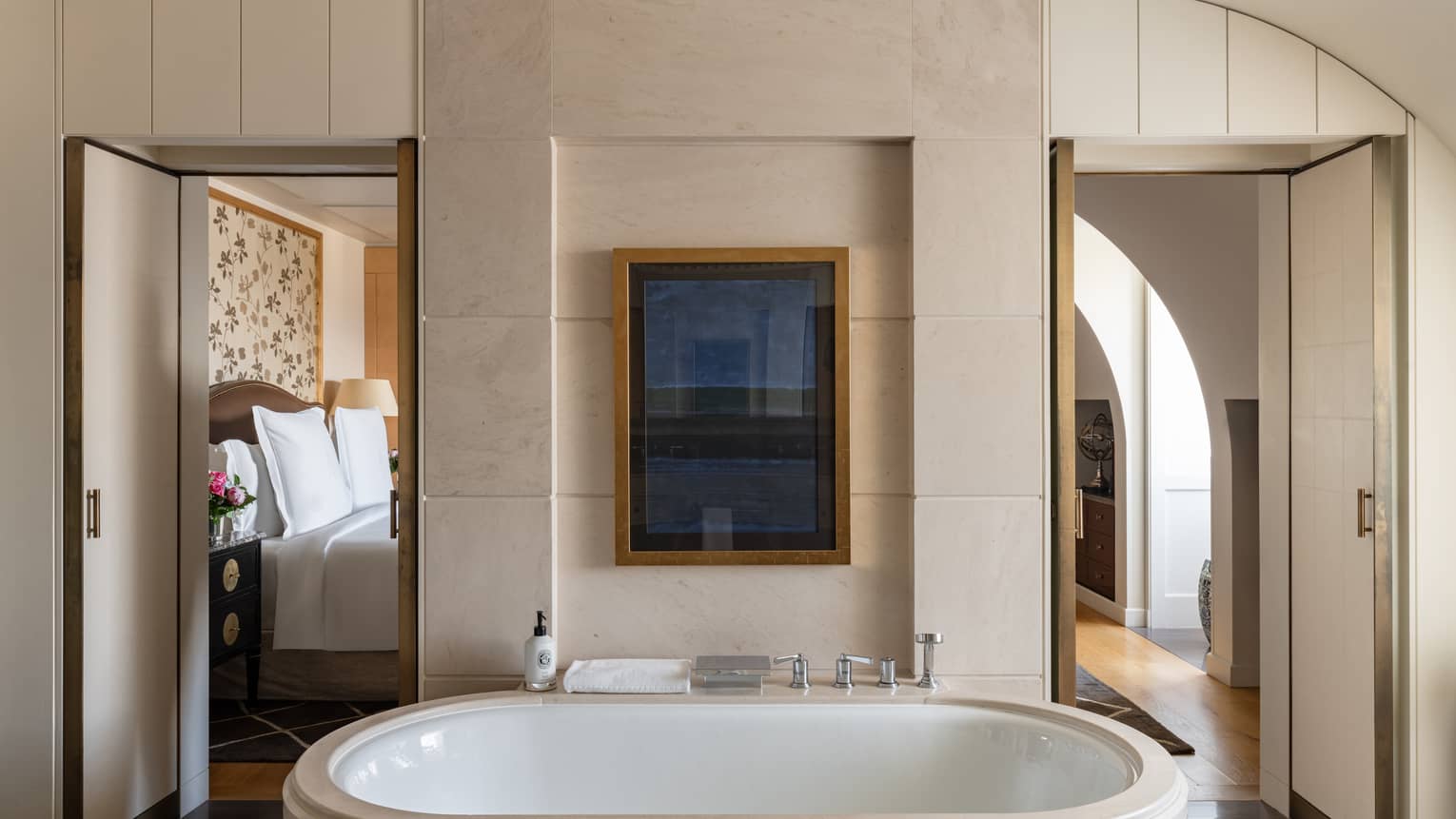 Luxury hotel bathroom with a soaking tub, minimalist décor and a framed artwork above, offering views into the bedroom with floral headboard design