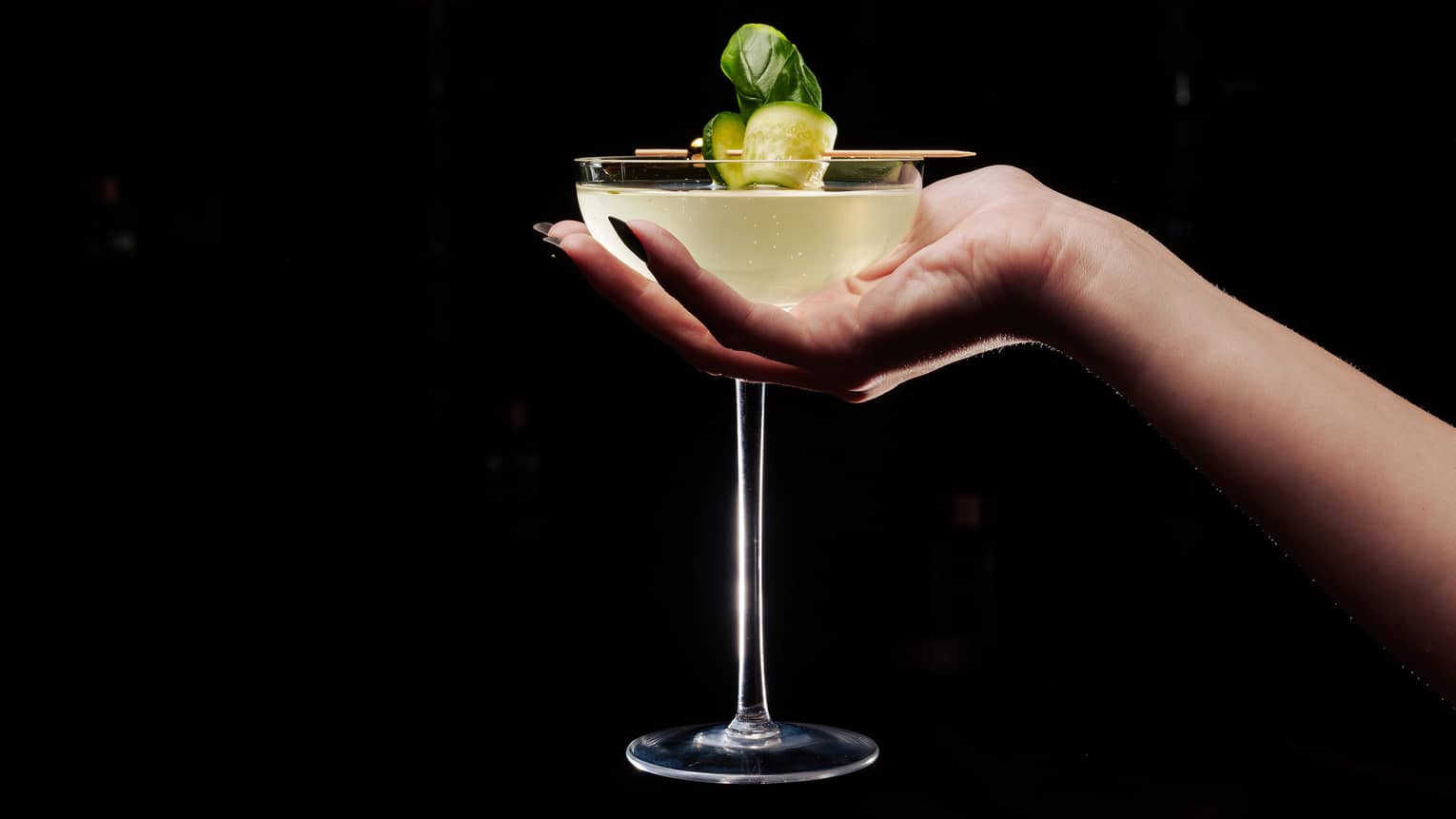 A hand holding a coupe cocktail glass filled with a yellow liquid and garnished with a curled piece of cucumber on a toothpick