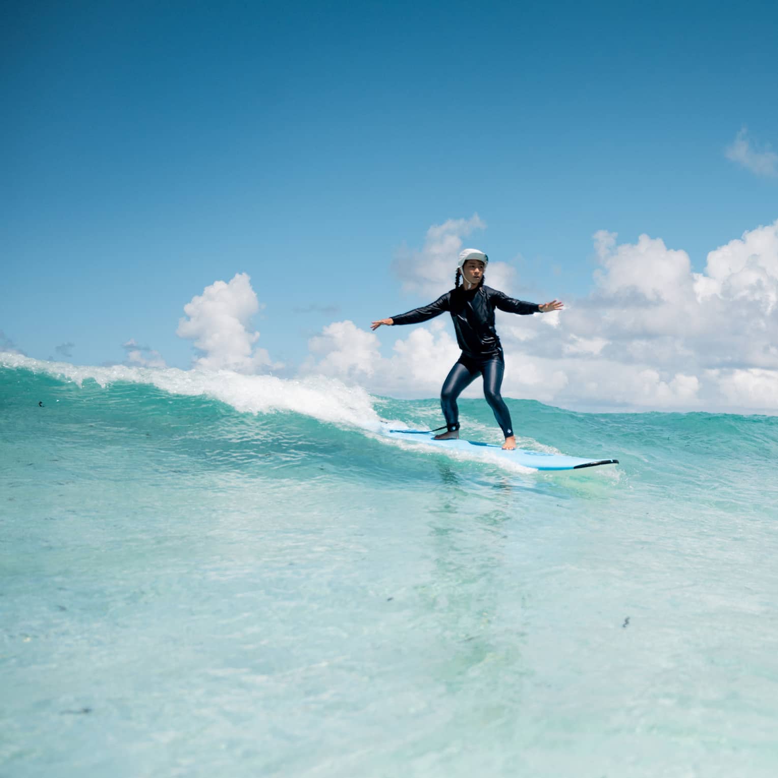 A wetsuit-clad surfer, arms outstretched, rides a turquoise wave under an expansive blue sky peppered with cumulus clouds.