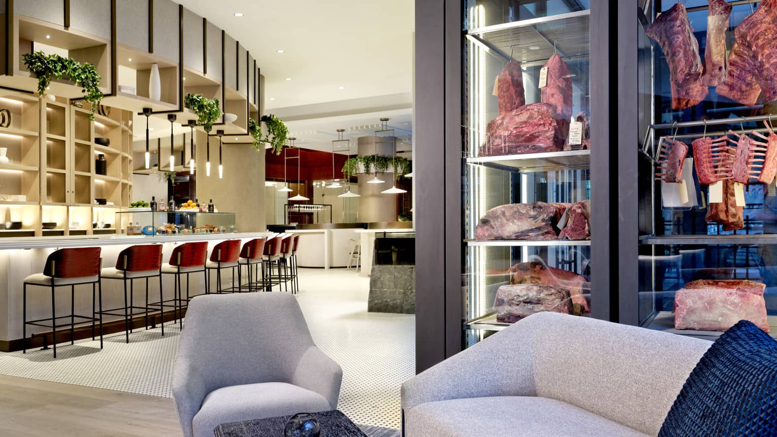 A lounge with grey chairs and a bar area with red stools and high shelves next to hanging meat in glass shelves ready for grilling.