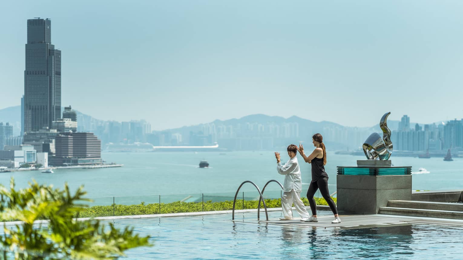 Two women in tai chi pose on platform on outdoor swimming pool overlooking Hong Kong waterfront