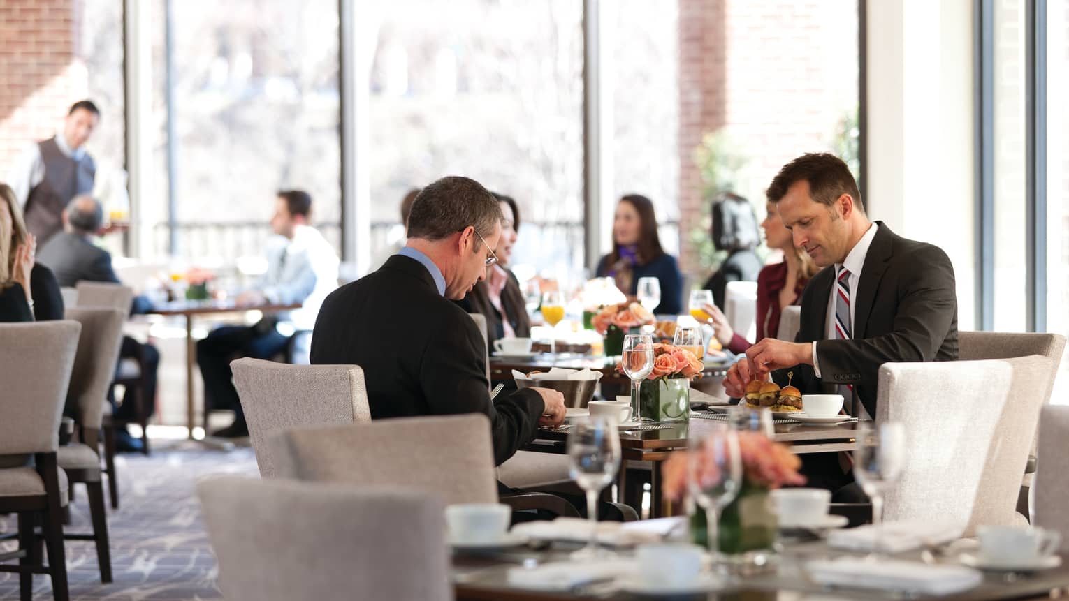 Men wearing suits dining around table in sunny Seasons restaurant, busy dining room in background
