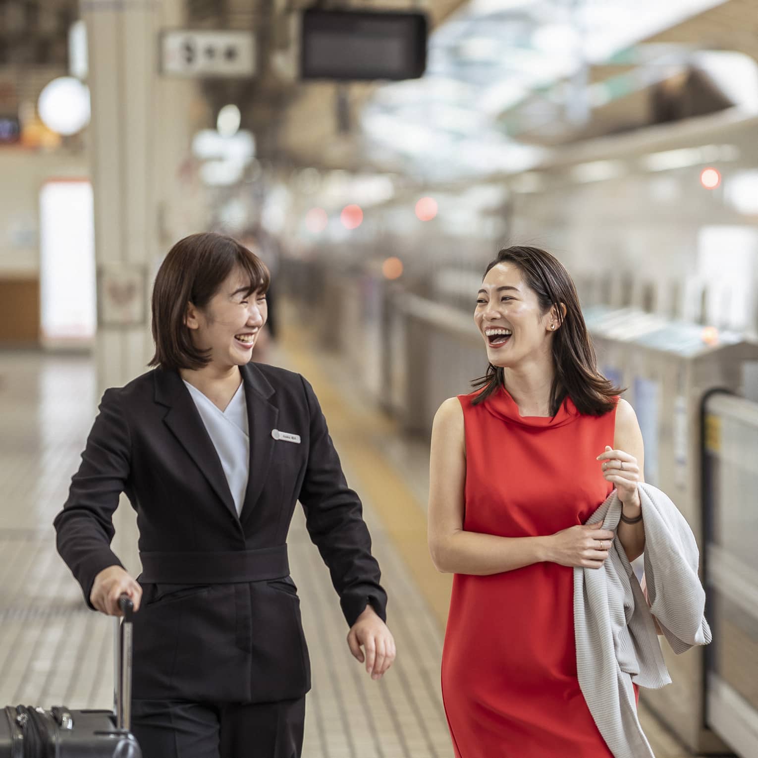 A Four Seasons employee holds a traveller's suitcase; laughing, they walk along the platform beside an N700 high-speed train.