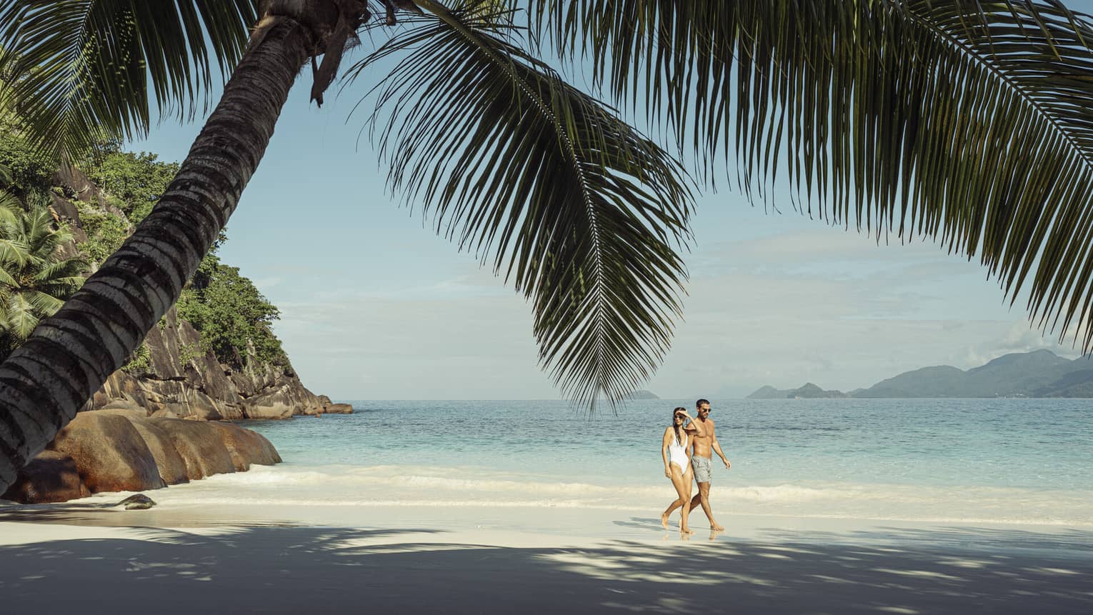 A couple walks along a beach shaded by a large palm tree