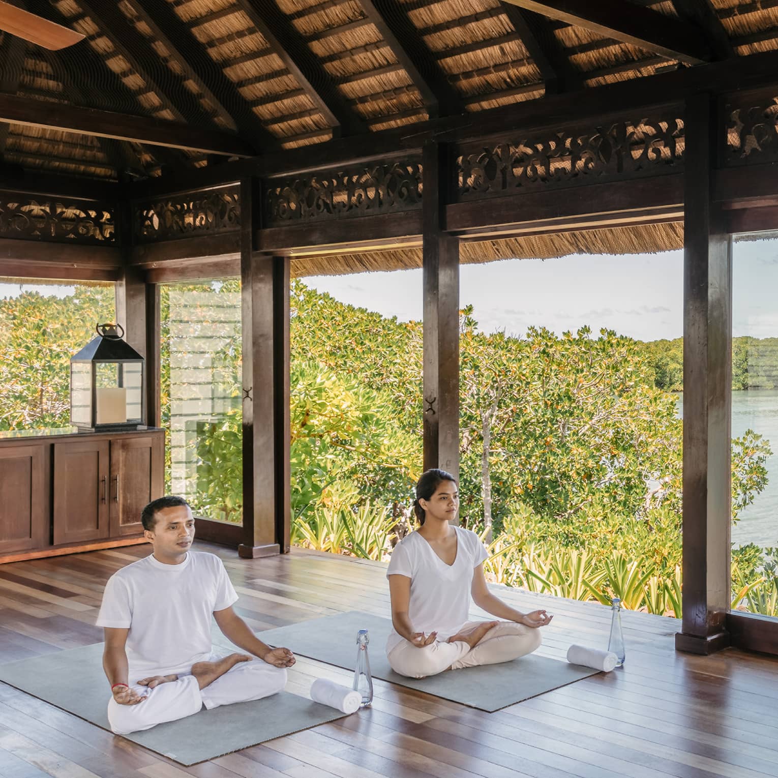 Two people wearing white seated in yoga poses in open-air spa yoga pavilion by beach