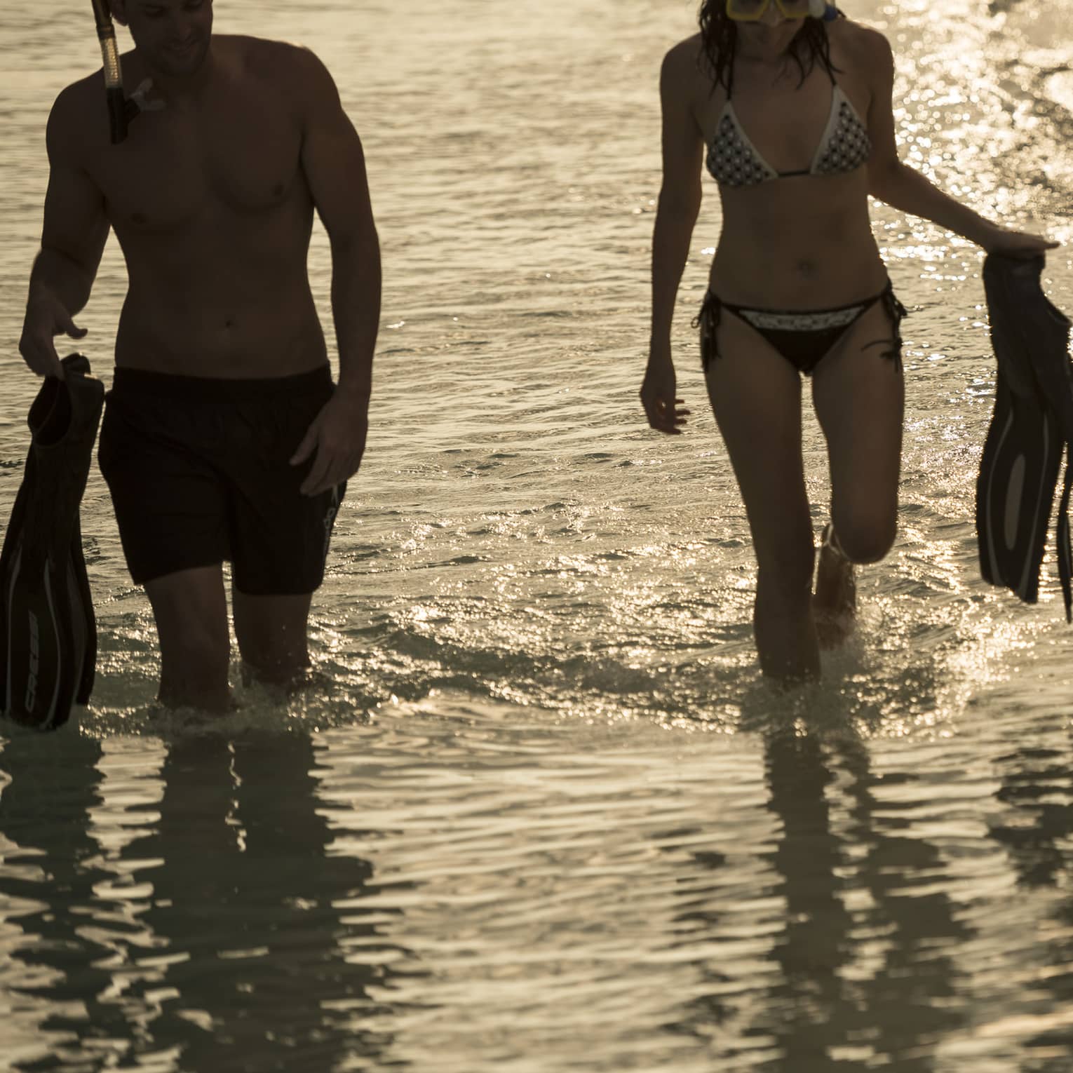 Two people in swimsuits and snorkel gear hold fins as they walk along the water's edge, sun reflecting off gentle waves.