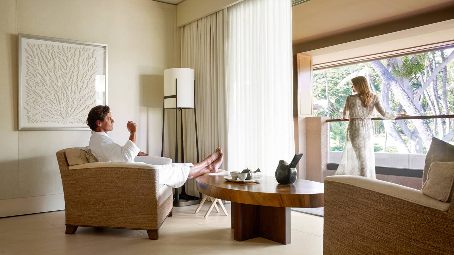 Man in white bathrobe kicks back in armchair in Spa room, woman in sheer robe stands at sunny window