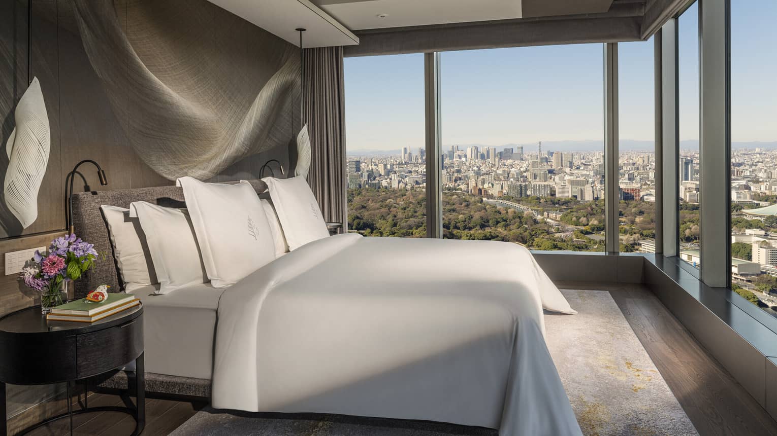 A gray upholstered bed, wooden night stand and modern light fixture in a room with stunning city views