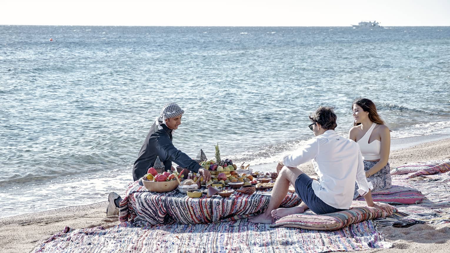 Three people sit on a beach around a low table set with a luxurious array of fruit, bread, and clay dishes full of food.