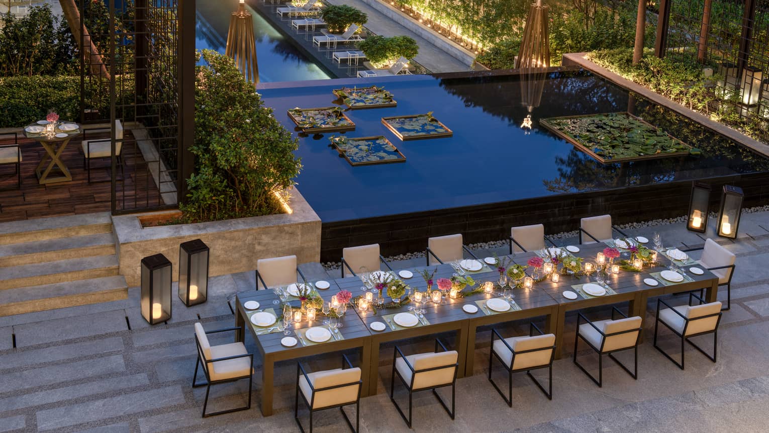 Outdoor terrace with long rectangular table and 12 chairs, nearby lotus pond and foliage at dusk